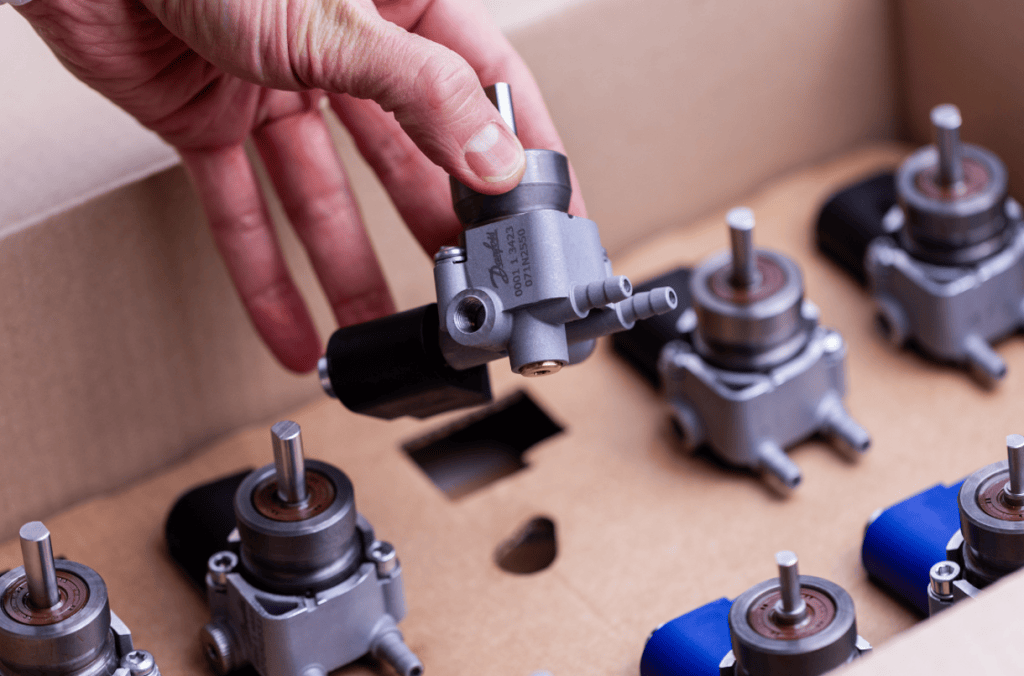 Käsi nostaa Danfoss-öljypumpun pahvilaatikosta, jossa on useita pumppuja.